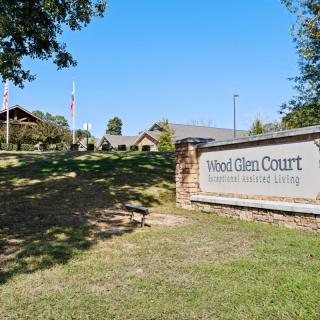 A sign for Hood Glen Court, an assisted living facility, with buildings and a landscaped area in the background.