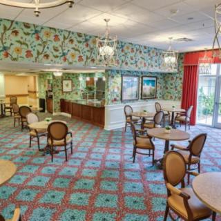 A well-lit dining area with multiple tables and chairs, featuring floral wallpaper and chandeliers.