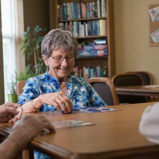 An elderly woman is smiling while playing cards at a table in a cozy room with bookshelves.