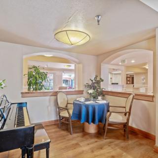 A cozy dining area with a piano, featuring a small table set for two and decorated with plants.