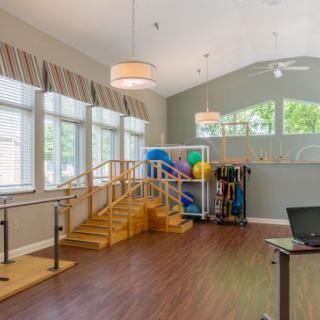 A bright physical therapy room featuring wooden stairs, parallel bars, and various rehabilitation equipment.