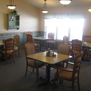 An empty dining room with multiple tables and chairs, featuring large windows and ceiling lighting.