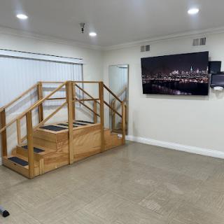 A spacious indoor room featuring a wooden stair structure, a large TV displaying a city skyline, and a whiteboard, suitable for therapy or training sessions.