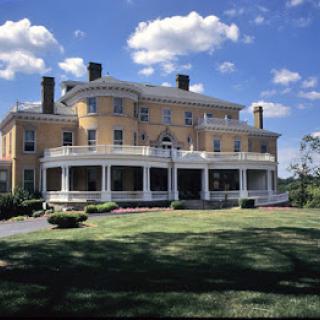 A historic mansion with a wraparound porch set against a bright blue sky with scattered clouds.