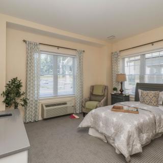 A cozy bedroom with a neatly made bed, two large windows with patterned curtains, a comfortable chair, and modern decor.