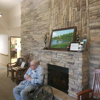 An elderly person in a wheelchair sits near a fireplace adorned with decorations and a painting on the mantel.