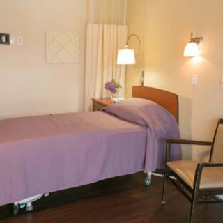 A cozy hospital room featuring a single bed with a purple blanket, a chair, and warm lighting.