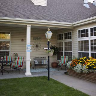 A cozy patio with outdoor furniture and blooming flowers, adjacent to a house with large windows.