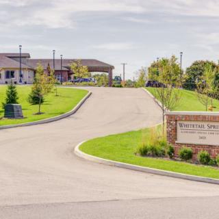 A view of a driveway leading to a building surrounded by greenery, with a sign at the entrance.