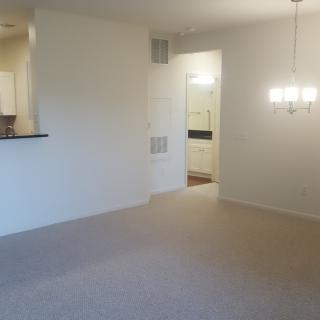 An empty apartment living room with a dining area, featuring a carpeted floor and a chandelier.