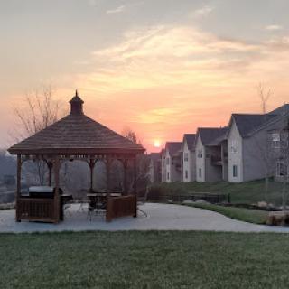 A serene sunset view featuring a gazebo, surrounded by lush grass and residential buildings.