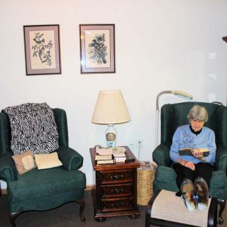 An elderly woman reading in a cozy living room with armchairs and framed art on the walls.