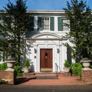A classic two-story house with a prominent wooden door, surrounded by greenery and tall trees.