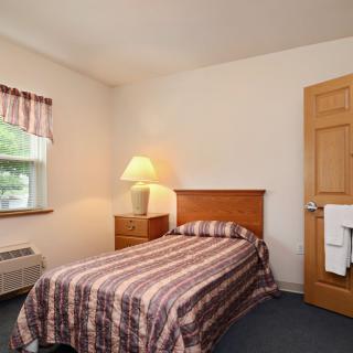 A cozy bedroom with a single bed, nightstand, and lamp beside a window.