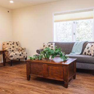 A cozy living room with a sofa, armchair, and wooden coffee table, decorated with plants and featuring natural light from a large window.