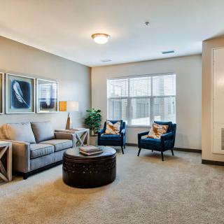 A cozy living room with a sofa, two armchairs, and a round coffee table, featuring artwork on the wall and natural light from the window.