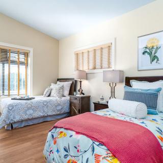 A well-lit bedroom featuring two neatly made beds with colorful bedding and decorative pillows, complemented by bedside tables and artwork on the walls.