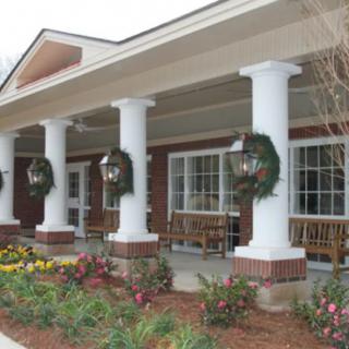 A brick building with a decorated porch and white columns, featuring wreaths and landscaped flowers.