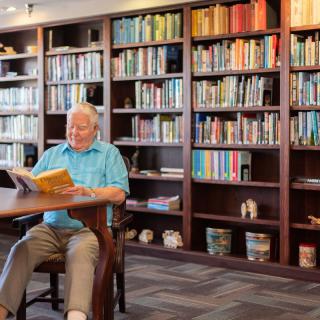 A senior man reading a book in a library with bookshelves filled with various books.