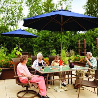 A group of seniors enjoying a relaxed afternoon on an outdoor patio, surrounded by greenery and shaded by blue umbrellas.