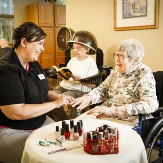 A caregiver provides a manicure to an elderly woman in a senior care setting, while another woman reads under a hair dryer.