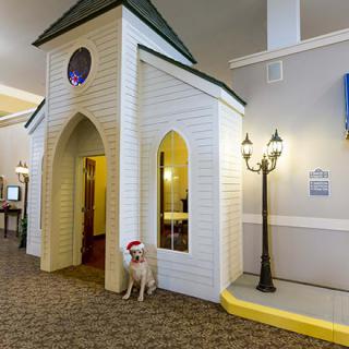 An indoor setting featuring a dog sitting beside a small building replica with Gothic-style windows and a lamp post.