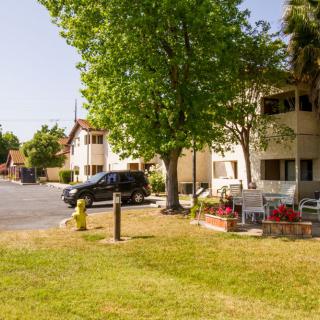 A sunny day in a residential area featuring apartment buildings, a parked car, and a grassy lawn with trees.