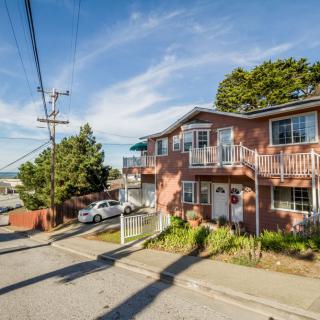 A two-story suburban house with balconies, situated on a quiet street with parked cars and surrounded by trees under a clear blue sky.