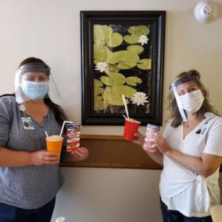 Two people wearing masks and face shields hold drinks in front of a framed piece of artwork indoors.