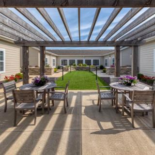A sunny patio with a pergola covering tables and chairs, overlooking a well-maintained garden and lawn.