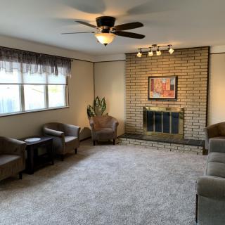 A cozy living room featuring a brick fireplace, plush carpet, and comfortable seating under a ceiling fan.