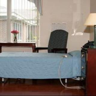 A hospital room with a neatly made bed, an armchair, and a table with flowers by the window.