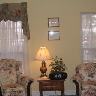 A cozy living room setup with two armchairs, a side table holding a lamp and plant, and decorated with sheer curtains.