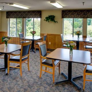 A cozy dining room with neatly arranged tables and chairs, adorned with floral centerpieces near large windows.