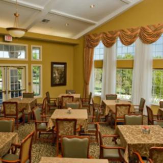 A well-lit dining room with tables and chairs arranged neatly, featuring large windows with curtains.