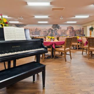 A dining area with a piano and music stand, surrounded by tables and chairs, featuring a mural on the wall.