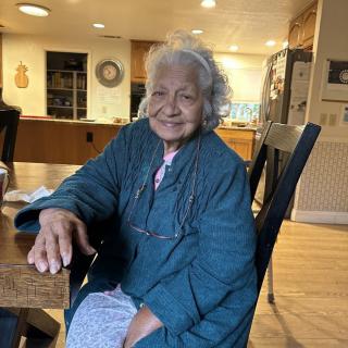 An elderly woman smiling while sitting at a table in a cozy kitchen.
