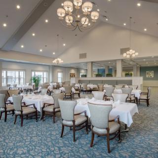 A spacious and elegant dining area featuring round tables with white tablecloths, upholstered chairs, and chandeliers hanging from the ceiling.