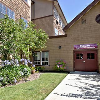The image shows the exterior entrance of a building with landscaped gardens, including flower beds and trees, along a paved pathway.