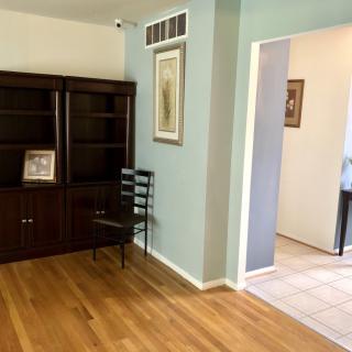 A well-lit living room with hardwood floors, showcasing a dark wooden bookshelf and a chair against a light blue wall adorned with artwork.