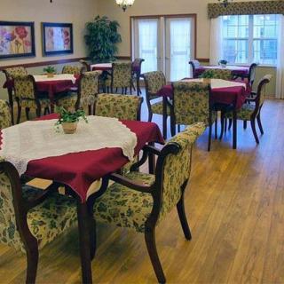 A cozy dining room with elegantly set tables and chairs, featuring floral decorations and wood flooring.