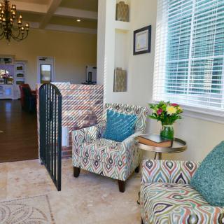 A cozy living room featuring patterned armchairs, a small round coffee table with a vase of flowers, and natural light streaming through the window blinds.