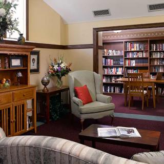 A cozy library reading room with an armchair, wooden table, and bookshelves filled with books.