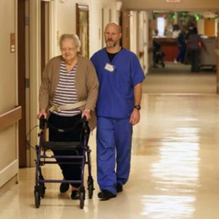 A caregiver assists an elderly person using a walker in a nursing home hallway.