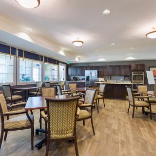 A spacious dining and kitchen area with multiple tables and chairs, illuminated by ceiling lights and natural light from large windows.