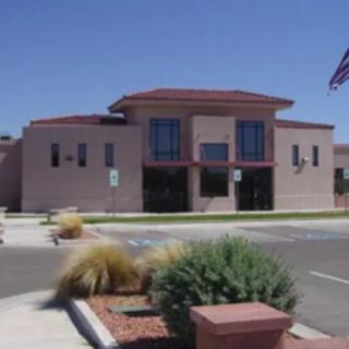A modern building with an American flag in front, surrounded by a parking lot and desert landscaping on a sunny day.