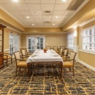 A well-lit dining room with a long table and chairs, featuring large windows and patterned carpet.