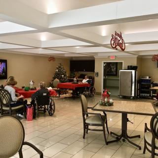 A cozy nursing home interior with decorated tables and seniors enjoying a TV program.