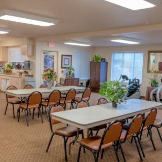 A community dining area with tables and chairs, decorated with indoor plants, under bright lighting.