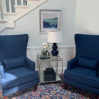A cozy living room corner featuring two blue armchairs, a stylish side table with a lamp, and a piece of artwork on the wall.